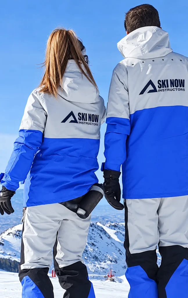 Dos instructores de Ski Now Instructors de espaldas, con uniformes azules y blancos, frente a las montañas nevadas de Sierra Nevada.