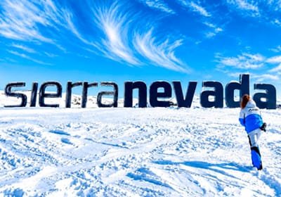 Cartel de Sierra Nevada sobre un paisaje de montañas nevadas, con una persona vestida de azul a la derecha.